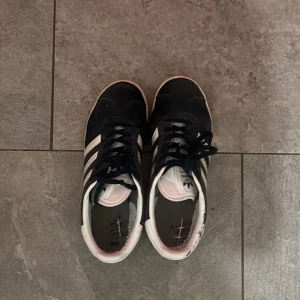 Adidas Gazelle sneakers marinblå - Säljer ett par klassiska Adidas Gazelle sneakers i marinblå mocka med vita detaljer och snörning. Skorna har de ikoniska tre ränderna på sidan, rund tå och platt sula med mönstrad undersida. Perfekt för dig som gillar retrostil och streetwear.