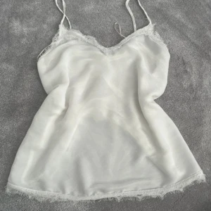 Vit spetskantad linnetopp från Zara - Säljer en vit linnetopp från Zara med tunna axelband och v-ringad hals. Toppen har transparent material med diskret prickmönster och spetsdetaljer längs halsringning och nederkant. Perfekt för att skapa en chic och feminin look.