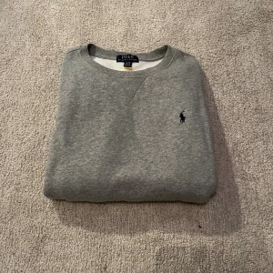 Grå sweatshirt från Polo Ralph Lauren XL - Grå sweatshirt från Polo Ralph Lauren i storlek XL. (Barn storlek) 18-20 passar typ 170 cm. Klassisk rund hals och broderad marinblå logga på bröstet. Mjuk insida och ribbade muddar vid hals, ärmslut och nederkant. Perfekt för chill dagar och enkel att matcha med jeans eller joggers.