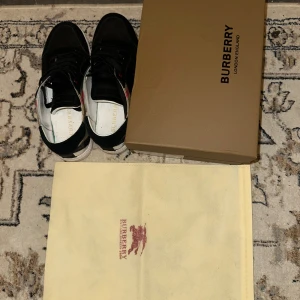 Burberry  - Snygga svarta sneakers från Burberry med klassiskt beige och svart rutmönster på sidorna, röda detaljer vid snörningen och vit platt sula. Skorna har ovandel i mocka och textil, samt svarta skosnören. Perfekta för dig som gillar stilrena och lyxiga sneakers. Mycket bra skicka