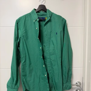 Grön skjorta från Polo Ralph Lauren - Snygg grön skjorta från Polo Ralph Lauren i bomull med klassisk button-down krage och broderad logga på bröstet. Skjortan har lång ärm, vita knappar och custom fit-passform. Perfekt för dig som gillar stilrena och tidlösa plagg.