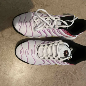 Vita Nike sneakers med lila detaljer - Säljer ett par vita Nike sneakers med lila vågiga mönster och svarta, röda samt gula detaljer. Skorna har snörning och en sportig vibe, perfekt för dig som gillar unika färgkombinationer. Ovandelen är i syntetmaterial och sulan är platt.