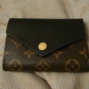 Monogram plånbok  - Snygg plånbok från i klassiskt monogrammönster med bruna och svarta toner. Plånboken har guldfärgade detaljer, tryckknapp och dragkedja. Tillverkad i canvas och har flera fack för kort och sedlar. Perfekt accessoar för dig som gillar lyxiga detaljer.