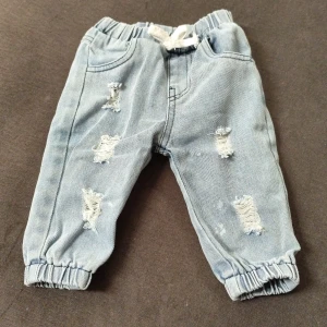 Ljusa jeansbyxor med slitningar från Shein - Säljer ett par ljusblå jeansbyxor från Shein i storlek 3-6 månader. Byxorna har coola slitningar framtill, resår i midjan och bensluten samt en snörning i midjan. Perfekt för en trendig look till de allra minsta.