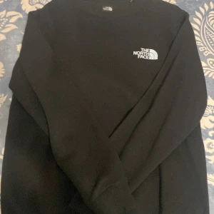 Svart sweatshirt från The North Face - Svart sweatshirt från The North Face i slim fit med klassisk logga på bröstet. Tröjan har rund hals och långa ärmar, tillverkad i mjuk bomullsmix som är skön mot huden. Perfekt för dig som gillar enkel och stilren streetwear.
