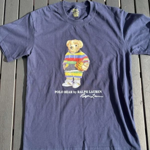 Blå Polo Bear t-shirt från Ralph Lauren - Snygg mörkblå t-shirt från Polo Ralph Lauren med ikonisk Polo Bear-tryck på bröstet. Björnen har en färgglad randig hoodie och håller en basketboll. T-shirten har rund hals, korta ärmar och är i slim fit. Perfekt för dig som gillar streetstyle och klassiska märken.