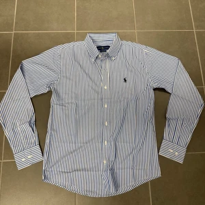 Blåvit randig skjorta från Ralph Lauren - Snygg blåvit randig skjorta från Ralph Lauren i klassisk passform. Skjortan har långa ärmar, knappar framtill och ikoniska Polo-loggan broderad på bröstet. Tillverkad i mjuk bomull och har en stilren krage med knappdetaljer.