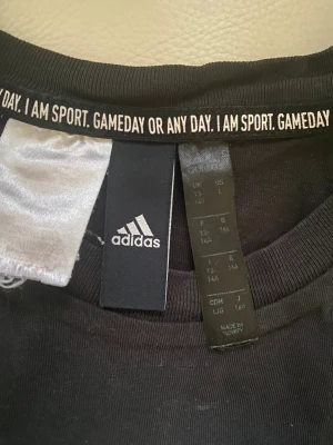 Svart Adidas t-shirt med logga, L - Svart t-shirt från Adidas med stor vit logga framtill. Klassisk rund halsringning och korta ärmar. Tillverkad i mjuk bomull, perfekt för en sportig och avslappnad stil. Snygg detalj med text på insidan av kragen. Det står L i den, men den känns mindre.. (om jag ska vara ärlig) L och M blir bra