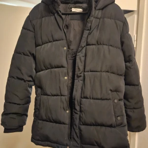 Svart pufferjacka från MANGO med huva - Svart pufferjacka från MANGO  i storlek S. Jackan har en skön huva, långa ärmar och praktiska fickor med tryckknappar. Den är quiltad och har dragkedja framtill. Perfekt för kalla dagar när du vill hålla dig varm och se chill ut.