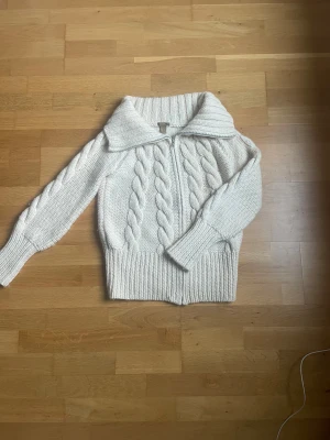 Vit stickad kofta från H&M med dragkedja - Säljer en vit stickad kofta från H&M i storlek S. Koftan har grova flätstickade detaljer framtill, bred ribbad krage och muddar samt dragkedja. Säljer pga fel storlek 😇