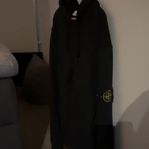 Svart hoodie från Stone Island - Svart hoodie från Stone Island med klassisk logotyp-patch på ärmen. Tröjan har huva med snörning och lång ärm. Perfekt för dig som gillar streetwear och vill ha en stilren look.