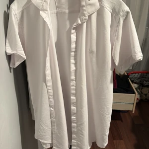 Vit kortärmad skjorta från Valentino - Säljer en stilren vit skjorta från Valentino med korta ärmar, klassisk krage och diskret broderad logga på bröstet. Skjortan har snygga guldfärgade detaljer vid kragen och är tillverkad i mjuk bomull. Perfekt för dig som gillar clean och exklusiv stil.