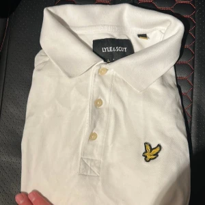Vit pikétröja från Lyle & Scott - Vit pikétröja från Lyle & Scott med klassisk krage och tre knappar framtill. Tröjan har det ikoniska broderade gula fågelmärket på bröstet. Tillverkad i mjuk bomull som känns skön mot huden och passar perfekt till jeans eller shorts.