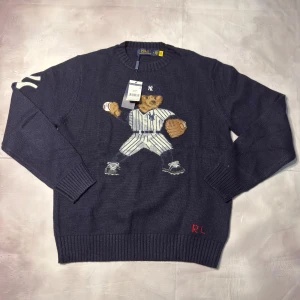 Mörkblå stickad tröja Polo Ralph Lauren - Mörkblå stickad tröja från Polo Ralph Lauren med broderad nalle i New York Yankees-baseballoutfit på framsidan. Rund hals, ribbade muddar och RL-broderi i rött vid nederkanten. Perfekt för dig som gillar sportiga och unika detaljer.