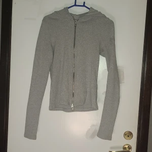 Grå hoodie med dragkedja från Weekday - Säljer en stilren grå hoodie från Weekday i storlek S. Tröjan är ribbad, har lång ärm, huva och hel dragkedja framtill. Materialet är mjuk bomull med lite stretch, perfekt för chill dagar. Passformen är normal men får även plats på en som har storlek Xs