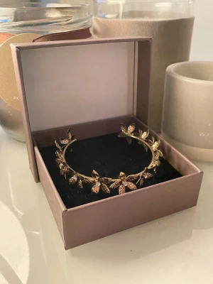 Guldfärgat armband med blommor - Superfint guldfärgat armband med detaljerade blommor och små glittrande stenar i mitten av varje blomma. Armbandet har en öppen design och kommer i en snygg ask, perfekt som present eller till dig själv om du vill sticka ut lite extra.