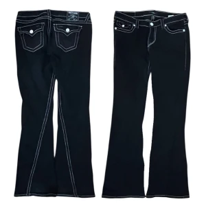 True Religion Joey Low Rise Flare jeans - Svarta jeans från True Religion, modell Joey Low Rise Flare. Snygga kontrastsömmar i vitt och rött, samt ikoniska ficklock med silverfärgade knappar bak. Låg midja och utsvängda ben ger en cool siluett. Perfekt för dig som gillar statement-jeans med detaljer 🖤🌹
