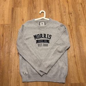 Morris Crewneck - Snygg grå stickad tröja från Morris med broderad text på bröstet. Tröjan har rund halsringning, ribbade muddar och klassisk collegestil. Perfekt för dig som gillar en stilren och sportig look.