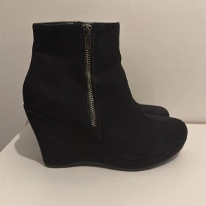 Svarta kilklacksstövletter från Graceland - Snygga svarta stövletter från Graceland med kilklack och rund tå. Skorna har en dragkedja på sidan för enkel på- och avtagning. Tillverkade i mocka-look som ger en mjuk och stilren känsla. Perfekta för dig som vill ha höjd men ändå stabilitet.