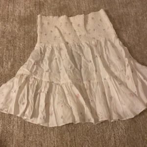 Vit blommig volangkjol - Söt vit kjol med små blommor i rosa och blå toner. Kjolen har volanger och en bred, ribbad midja som ger en skön passform. Perfekt för dig som gillar romantisk och feminin stil. Materialet känns lätt och luftigt, perfekt för varmare dagar.