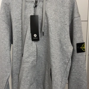 Grå hoodie från Stone Island med hel zip - Säljer en grå hoodie från Stone Island med klassisk logopatch på ärmen. Tröjan har huva med snörning, half zip framtill och en stor magficka. Materialet är mjuk bomull och passformen är loose, perfekt för chill dagar.