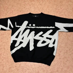 Svart Stüssy stickad tröja med logga - Säljer en svart stickad tröja från Stüssy med stor vit logga i graffiti-stil över hela framsidan och baksidan. Tröjan har rund hals och långa ärmar, perfekt för dig som gillar streetwear och vill sticka ut med en fet design.