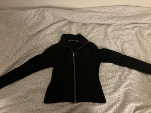 Svart ribbad kofta med dragkedja - Svart ribbad kofta med hel dragkedja framtill och hög krage. Tröjan har långärmad passform och är figurnära, vilket ger en snygg siluett. Perfekt för dig som gillar stilrena och enkla plagg med lite edge.