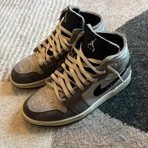 Nike Air Jordan 1 mid grå/svart - Säljer ett par Nike Air Jordan 1 High sneakers i grått och svart med vita detaljer. Skorna har klassisk snörning, perforerad tå och hög siluett. Materialet är en mix av mocka och syntet, med svart swoosh på sidan och svart yttersula. Perfekt för dig som gillar streetwear och ikoniska sneakers.