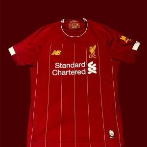 Liverpool FC matchtröja New Balance - Liverpool FC matchtröja från New Balance i röd färg med tunna vita ränder. Klassisk rund hals med vita detaljer och klubbmärke samt sponsortryck på bröstet. Tillverkad i lätt och ventilerande funktionsmaterial.