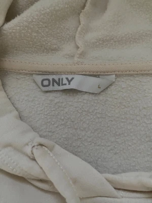 Beige hoodie från ONLY i storlek L - Säljer en beige hoodie från ONLY i storlek L. Tröjan har huva med snörning, midjekort och är långärmad. Materialet är mjukt och känns skönt mot huden. Enkel och stilren design utan mönster eller tryck.