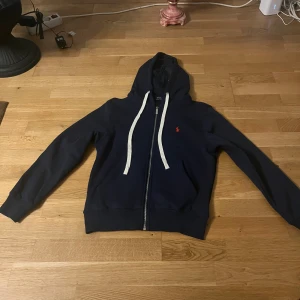 Marinblå hoodie från Polo Ralph Lauren - Snygg marinblå hoodie från Polo Ralph Lauren med vit snörning och dragkedja framtill. Tröjan har huva, långa ärmar och röd broderad logga på bröstet. Tillverkad i mjuk bomull för skön komfort. Perfekt för chill dagar och streetstyle.