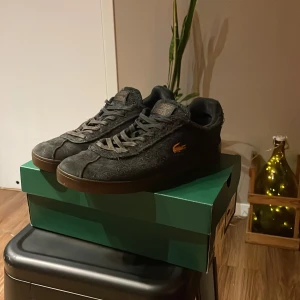 Svarta sneakers från Lacoste i mocka storlek 43 - Säljer ett par svarta Lacoste sneakers i mocka med bruna sulor och guldfärgad krokodillogga på sidan. Skorna har snörning och en klassisk låg profil. Perfekta för dig som gillar stilrena och sportiga skor med en twist.