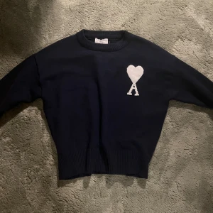 Mörkblå stickad tröja från Ami Paris - Säljer en mörkblå stickad tröja från Ami Alexandre Mattiussi med vit hjärtlogga på bröstet. Tröjan har rund hals, ribbade muddar och är långärmad. Perfekt för dig som gillar stilrena detaljer och vill ha något som sticker ut lite. Säljer pga för liten. Köpt här på plick så vet ej om den är äkta eller inte, men fick med tags och påse.