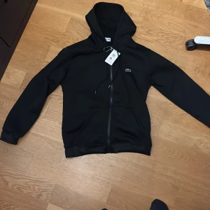 Helt ny Lacoste zip up hoodie  - Svart hoodie från Lacoste i klassisk stil med huva och snörning. Tröjan har dragkedja framtill, två fickor och det ikoniska krokodilloggan på bröstet. Perfekt för chill dagar och enkel att matcha med jeans eller joggers.
