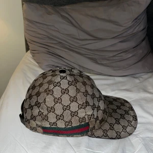 Gucci keps beige - Snygg keps från Gucci med klassiskt GG-monogram i brunt och beige. Kepsen har justerbart spänne i skinn baktill och en ikonisk grön-röd rand på sidan. Tillverkad i bomullscanvas med detaljer i skinn. Perfekt för dig som vill sticka ut med en lyxig accessoar.