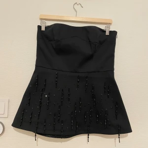 Svart axelbandslös topp med pärlor - Supersnygg svart axelbandslös topp. Toppen har en utsvängd kjol och är dekorerad med svarta pärlor och paljetter framtill för extra detalj. Perfekt för jultider och nyår! Använd fåtal gången och i bra skick. 