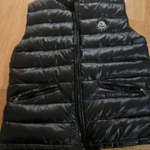 Svart dunväst från Moncler - Snygg svart dunväst från Moncler med quiltad design. Västen har två fickor med dragkedja framtill och klassisk Moncler-logga på bröstet. Perfekt att slänga över en hoodie för extra värme och stil.