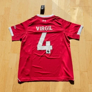 Liverpool Virgil #4 matchtröja Adidas storlek M - Liverpool FC fotbollströja med Virgil och nummer 4 på ryggen. Röd med vita detaljer, Adidas-logga och klubbmärke framtill. Kortärmad modell i lätt funktionsmaterial, med vita ränder på axlarna och sponsortryck på bröstet.