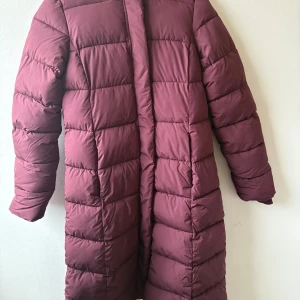 Vinröd lång pufferjacka från 157 - Säljer en lång och mysig vinröd pufferjacka från 157 med huva och dragkedja. Jackan har knappar framtill, två fickor med dragkedja och quiltad design. Perfekt för kalla dagar och har en loose passform som ger extra komfort. Ny utan prislapp  nypris 500:-