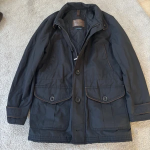 Mörkblå parkajacka från Massimo Dutti - Mörkblå parkajacka från Massimo Dutti med quiltat foder, stora fickor med lock och knappstängning samt dragkedja och knappar framtill. Jackan har bruna detaljer vid sömmarna och är långärmad med klassisk krage. Perfekt för höst och vinter.