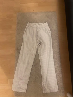 Randiga linnebyxor från 157 XS - Snygga ljusbeige linnebyxor från 157 i storlek XS. Byxorna har tunna vertikala ränder i vitt och beige, elastisk midja med snörning och raka ben. Perfekta för chill dagar och har en skön, luftig känsla tack vare linnematerialet.