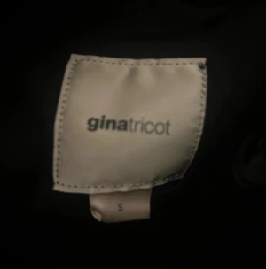 Svart kappa med fuskpäls från Gina Tricot - Svart kappa från Gina Tricot i storlek S med snygg fuskpäls på huvan och klassiska knappar framtill. Jackan har en rak passform och är tillverkad i ullmix, perfekt för kyliga dagar. Stilsäker look med fickor och mjukt material. En trasig knapp 