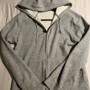 Grå hoodie från Polo Ralph Lauren - Snygg grå hoodie från Polo Ralph Lauren med dragkedja, huva och snörning. Tröjan har broderad logga på bröstet och är tillverkad i mjuk bomullsmix. Perfekt för chill dagar och enkel att matcha med jeans eller joggers.