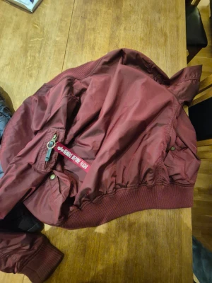 Vinröd bomberjacka från Alpha Industries - Säljer en vinröd bomberjacka från Alpha Industries med klassisk 'Remove Before Flight'-tag på ärmen. Jackan har orange foder, ribbade muddar och dragkedja framtill. Perfekt för dig som gillar streetwear och vill sticka ut med en ikonisk look.