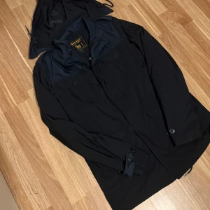 Svart fildejacket från Woolrich M/L - Svart parkajacka från Woolrich med avtagbar huva och knappar framtill. Jackan har två bröstfickor med lock och justerbara ärmslut med knapp. Materialet är slitstark polyester och jackan har en klassisk, rak passform. Perfekt för kyliga dagar och snygg till streetwear.