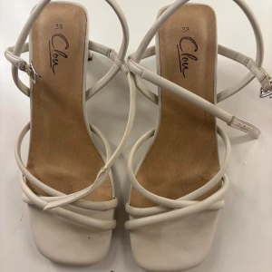 Beige sandaler med klack från Clou - Snygga beige sandaler från Clou med smala remmar och fyrkantig tå. Skorna har en elegant klack och justerbart spänne vid ankeln. Perfekt för dig som gillar stilrena och trendiga skor med en feminin touch.