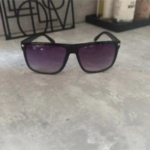 Två par retro solglasögon - Säljer två par solglasögon med klassisk fyrkantig form. Ena paret har svart båge med mörklila tonade glas, det andra har brun båge med brunt tonade glas. Båda är tillverkade i syntetmaterial och har en stilren vibe som passar perfekt till sommaren.