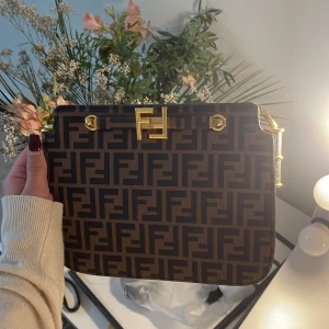 Fendi brun axelväska med FF-mönster - Säljer en brun axelväska från Fendi med ikoniskt FF-mönster i svart. Väskan har guldiga metalldetaljer och ett stilrent, fyrkantigt format. Insidan är fodrad i gult och har flera fack. Perfekt för dig som vill ha en statementväska med lyxig känsla. 