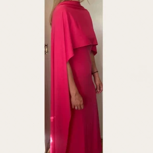 Ceriserosa långklänning med cape/sjal detalj - Oanvänd ceriserosa ärmlös långklänning. Kortärmad klänning med en lös rosa sjal som kan stylas hur man vill. Rund halsringning. I stolek M men passar även storlek S