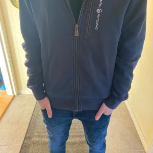 Mörkblå hoodie från Sail Racing - Mörkblå hoodie från Sail Racing med dragkedja framtill och logga på bröstet. Tröjan har huva, långa ärmar och är tillverkad i mjukt bomullsmaterial. Perfekt för chill dagar och har en clean, sportig look. Inte använd så många gånger 